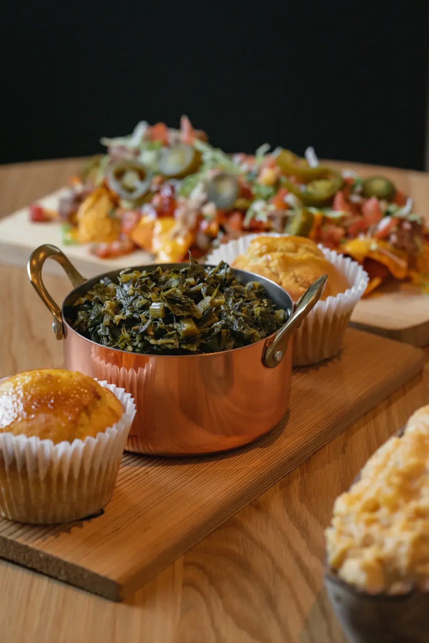 A catering spread of collard greens, cornbread muffins, and loaded nachos on wooden boards